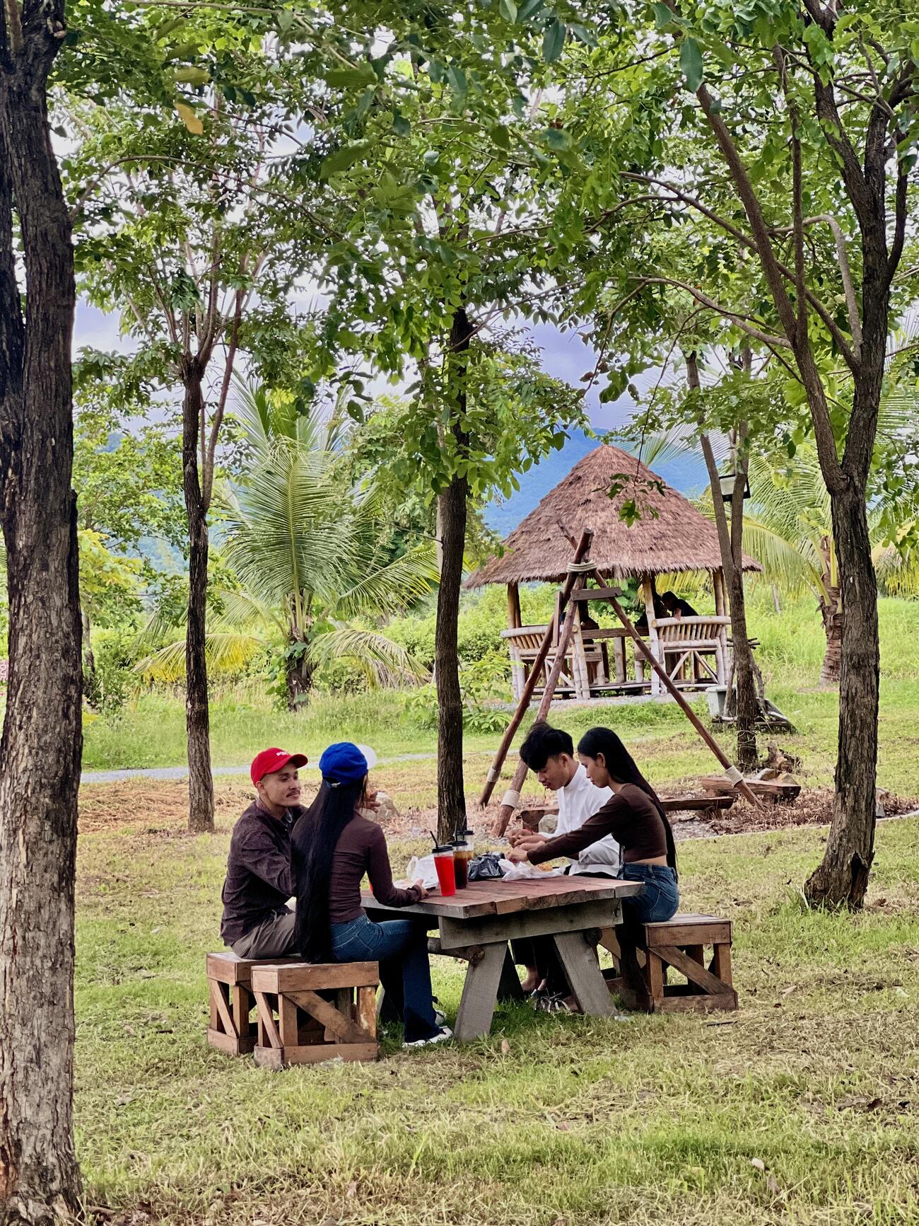 Friends gathered around a wooden table beside a thatched Kanhchos pavilion